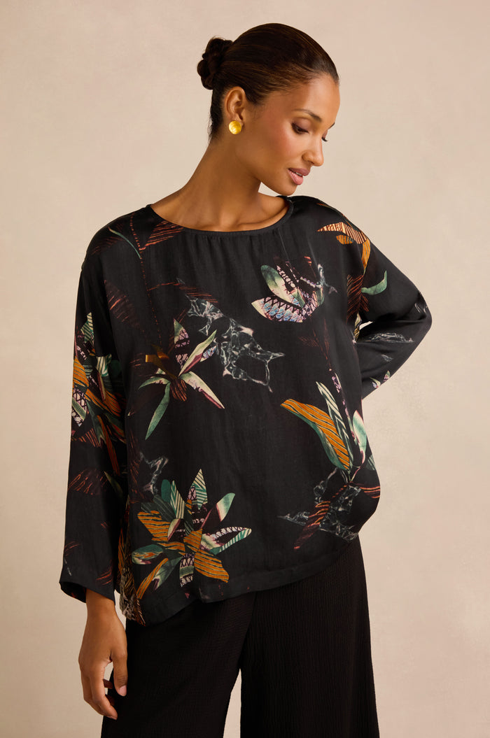 A woman wearing the Abstract Floral Boxy Top—featuring a loose fit and vibrant abstract patterns in black—paired with black pants and gold earrings, her hair styled in a neat bun as she looks down to her left against a plain background.