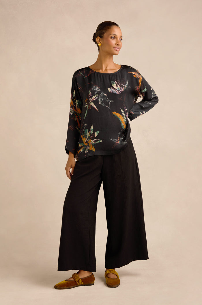 Wearing the Abstract Floral Boxy Top, a woman stands against a plain beige background in wide black pants and brown shoes, gazing to the side with one hand on her hip.