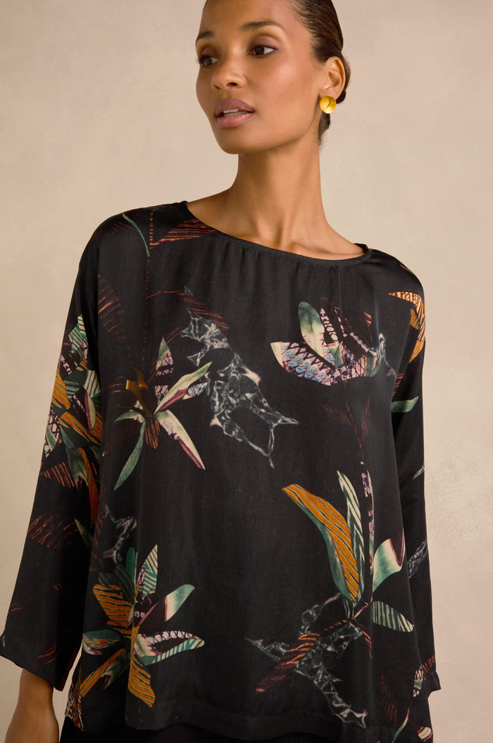 A woman is wearing the Abstract Floral Boxy Top featuring colorful patterns, paired with small yellow earrings, standing against a neutral background.