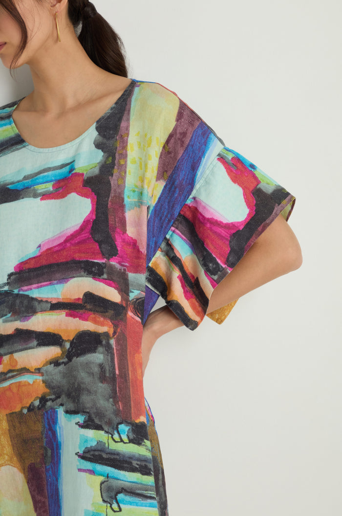 A woman stands against a plain background wearing the Abstract Print Easy Dress, featuring a relaxed silhouette and colorful abstract pattern, paired with black sandals.