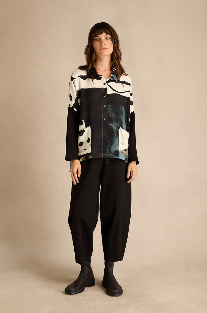 A woman stands against a neutral background wearing the Abstract Watercolour Shirt, paired with loose black pants and black boots.