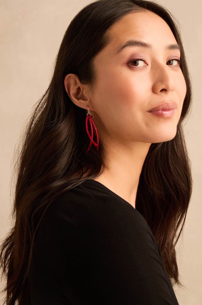 A woman with long dark hair wearing a black top showcases the Arc Earrings—striking red earrings with sterling silver hooks—as she looks at the camera against a plain beige background.