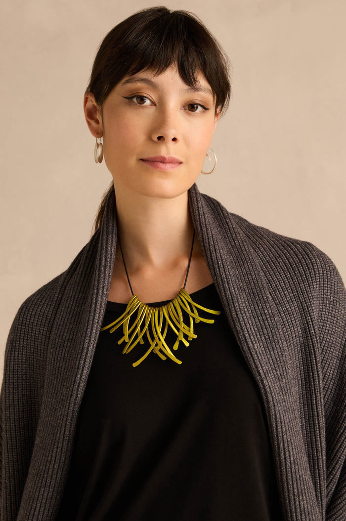 Model with straight dark hair wears a dark shawl, black top, and silver hoop earrings, accessorised with the Arc Necklace—a bold yellow statement piece crafted from anodised aluminium—creating an eye-catching example of contemporary jewellery, set against a neutral background.