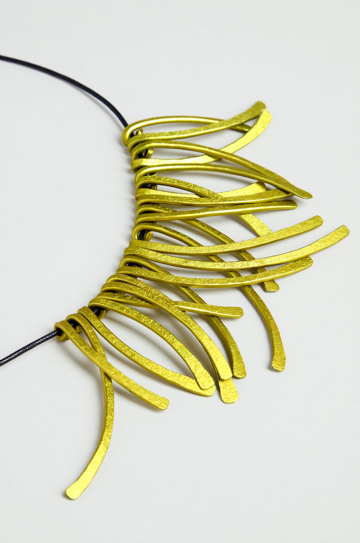 Introducing the Arc Necklace: a contemporary piece crafted from multiple curved, flat yellow anodised aluminium elements, artfully arranged in a striking fringe pattern along a black cord. Displayed on a light background, this bold yet elegant necklace embodies wearable art through its modern design.
