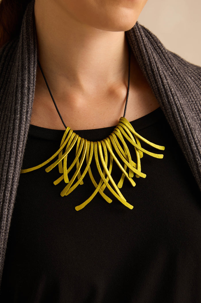 A person wearing a black top and gray cardigan, showcasing the Arc Necklace—an eye-catching piece of contemporary jewellery crafted from bold yellow anodised aluminium with multiple curved, tube-like elements that double as wearable art.