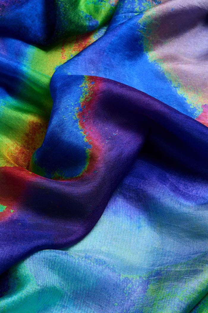 A close-up of the Aurora Veil Silk Scarf, featuring smooth, flowing pure silk with a vibrant abstract print in blue, green, red, and yellow.