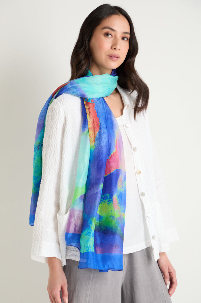 A woman in a textured white blouse and grey pants faces the camera, wearing the Aurora Veil Silk Scarf—pure silk with an abstract print in vibrant blue, green, and red—draped around her neck.