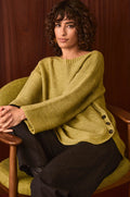 A woman with curly hair sits in a green chair against a wooden background, wearing the Avocado Ribbed Wool Blend Button Jumper—a cozy, loose olive knit with side buttons—paired with dark pants.