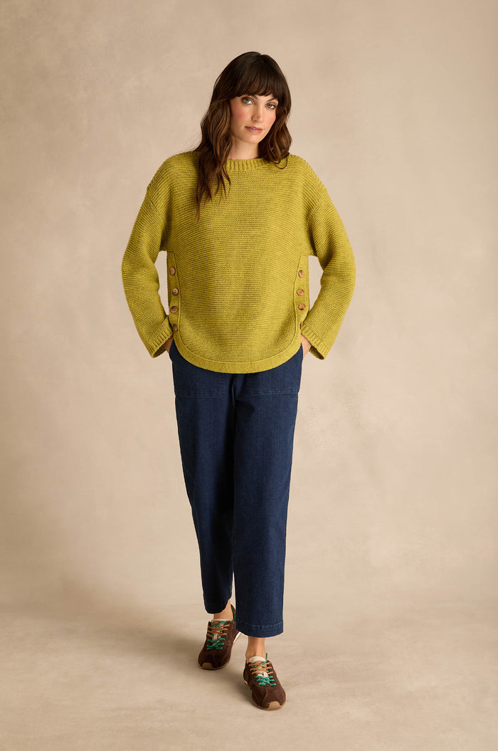 Woman wearing an Avocado Ribbed Wool Blend Button Jumper with blue cropped pants and brown shoes, standing against a beige background.