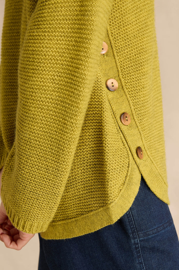 Person wearing the Avocado Ribbed Wool Blend Button Jumper, a textured yellow-green sweater with side button detail, paired with blue denim jeans.