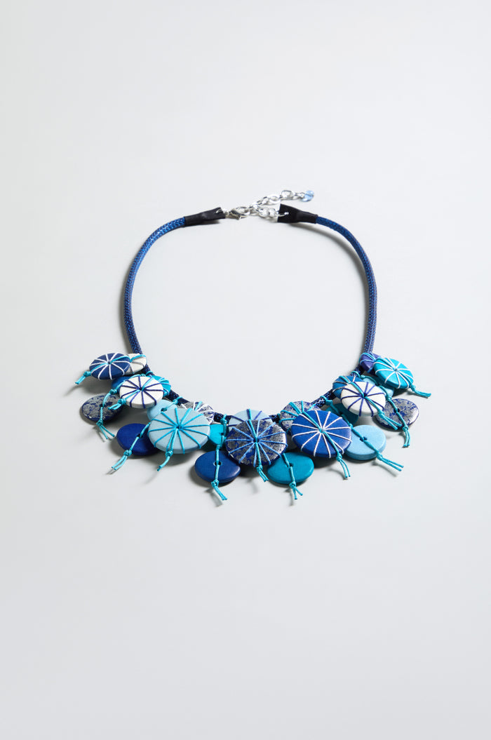 The Azure Short Disc Necklace features round, patterned blue and white beads with cord detailing. Adjustable for versatile style, it’s shown laid flat on a light grey background. Perfect for any occasion.
