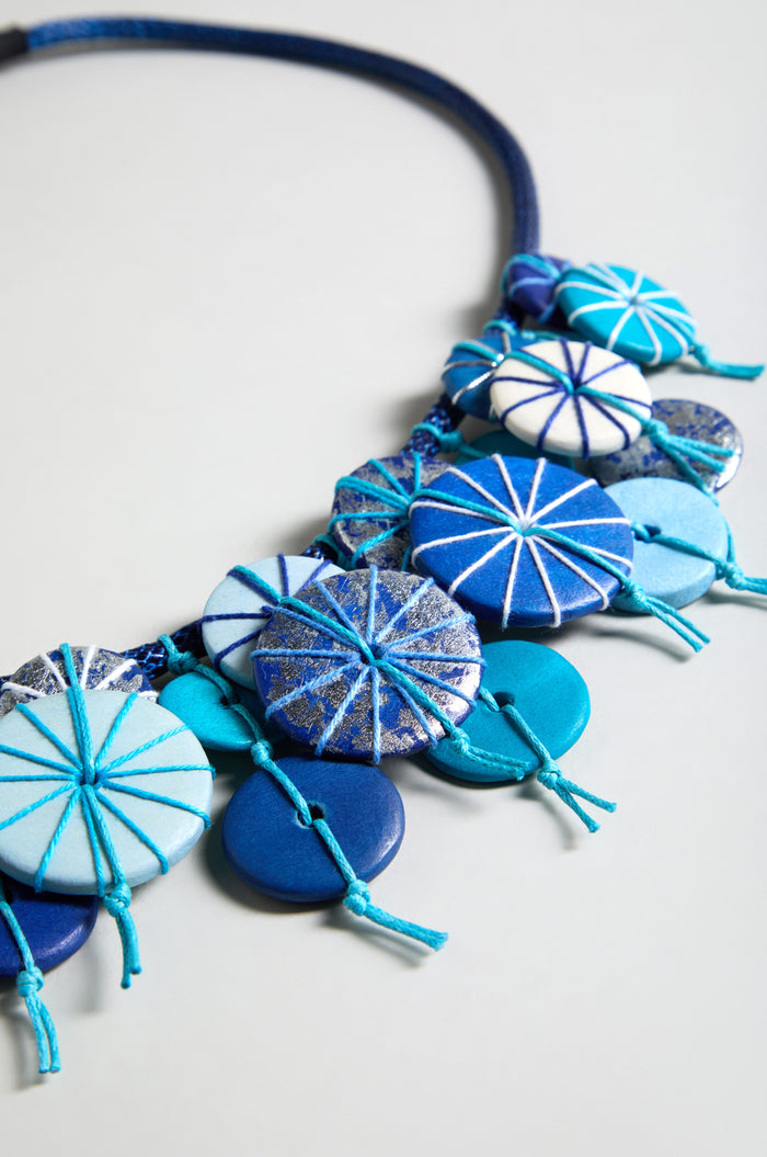 The Azure Short Disc Necklace is an adjustable blue necklace with round beads and wooden discs featuring geometric patterns and knotted string accents, shown against a light background.
