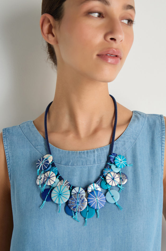 A woman in a sleeveless light blue top wears the Azure Short Disc Necklace—an adjustable blue necklace with circular, patterned beads.