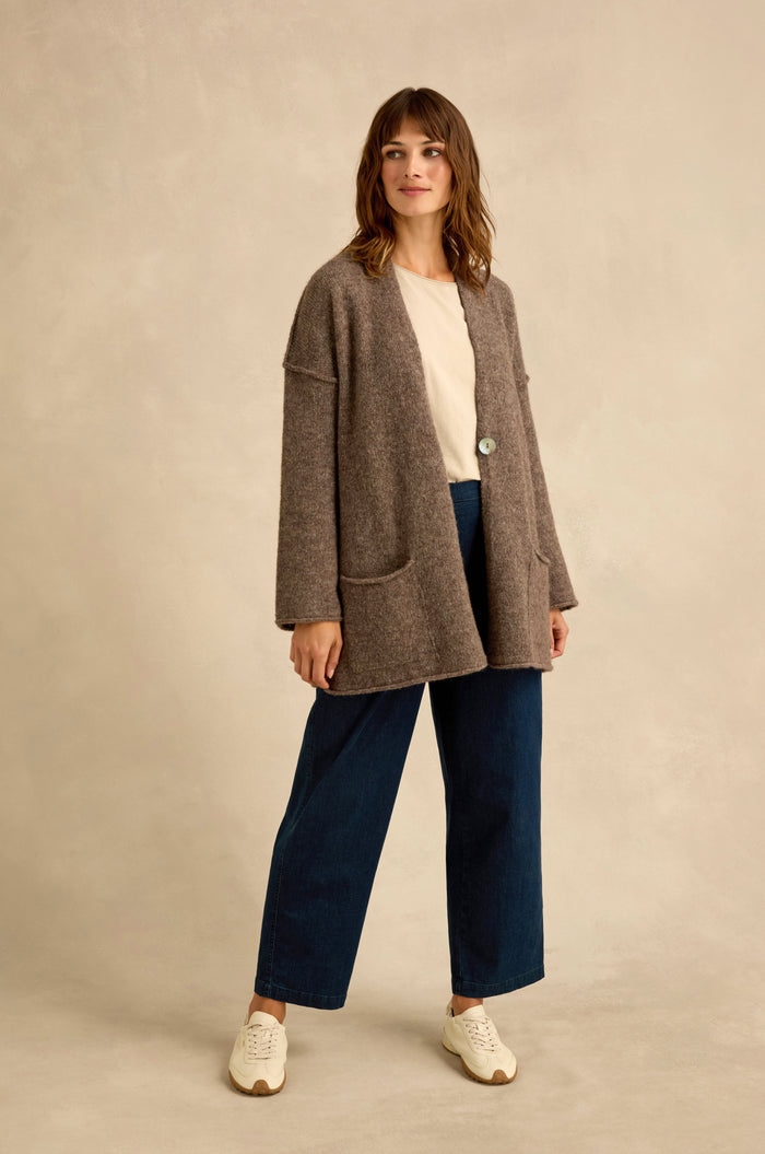 A woman stands against a beige background, wearing a beige top, wide-leg blue jeans, white sneakers, and the Bark Alpaca Blend Long Line Cardigan.