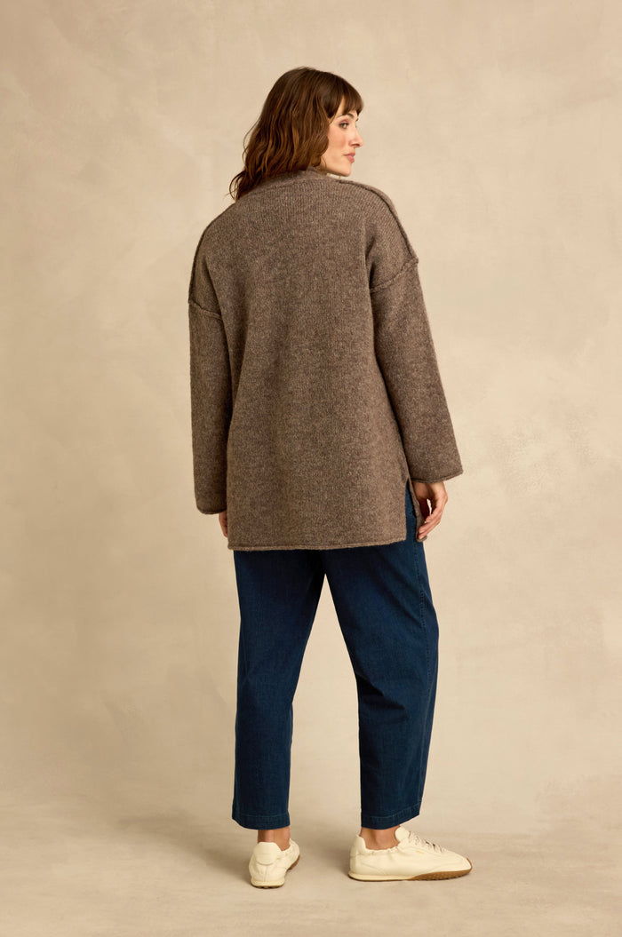 A woman stands facing away, wearing the Bark Alpaca Blend Long Line Cardigan with blue jeans and white sneakers against a neutral background.