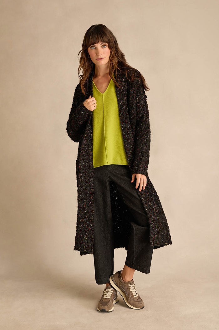 A woman stands in a studio against a neutral background, wearing the Black Boucle Knit Long Line Speckle Cardi layered over a lime green V-neck top, paired with black pants and beige sneakers.