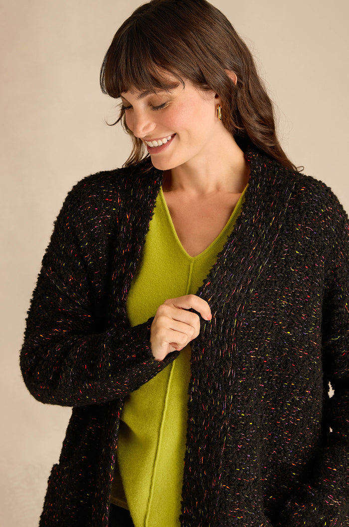 A woman with dark hair wears a bright green V-neck top and the Black Boucle Knit Long Line Speckle Cardi while looking down and smiling.