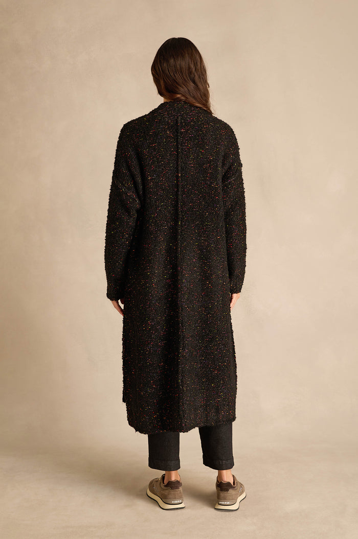 A person stands with their back to the camera, wearing the Black Boucle Knit Long Line Speckle Cardi, paired with dark pants and beige sneakers against a plain beige background.