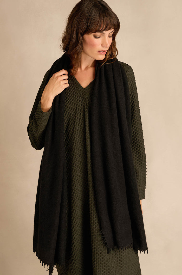 A woman stands against a neutral background, looking down as she holds the Black Soft Cashmere Scarf draped over her olive textured long-sleeve dress.