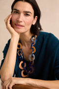 A woman with short dark hair wears a teal sweater and the Blue Stripe Wooden Resin Links Necklace—a striking statement piece featuring large, linked elements in brown, blue, and silver tones with bold resin and wood accents—as she sits with her hand resting on her face.