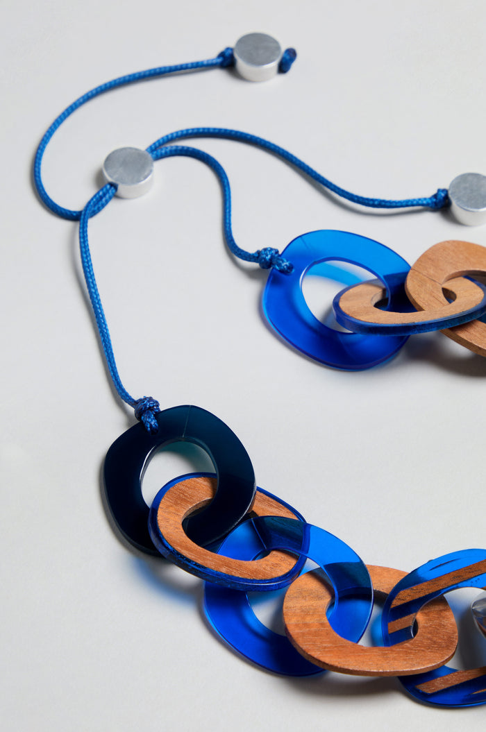 The Blue Stripe Wooden Resin Links Necklace features alternating blue plastic and wooden oval links, each with a striped design, strung on a blue cord with white circular end pieces, and is displayed against a light background.