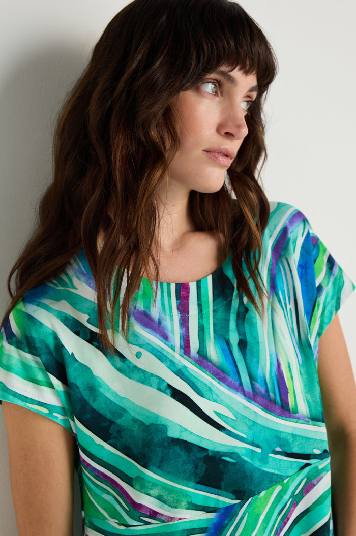 Woman with wavy brown hair looks to the side, wearing the Blurred Lines Swing Dress—a short-sleeved, stretch jersey dress featuring green, blue, and white abstract patterns.