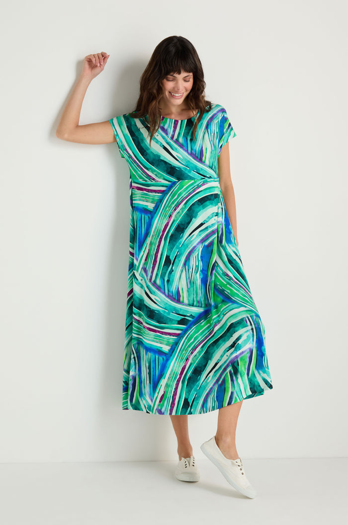 A woman stands against a white wall wearing the Blurred Lines Swing Dress, featuring a green, blue, and white abstract pattern in soft stretch jersey. The short-sleeve midi dress is styled with classic white sneakers.