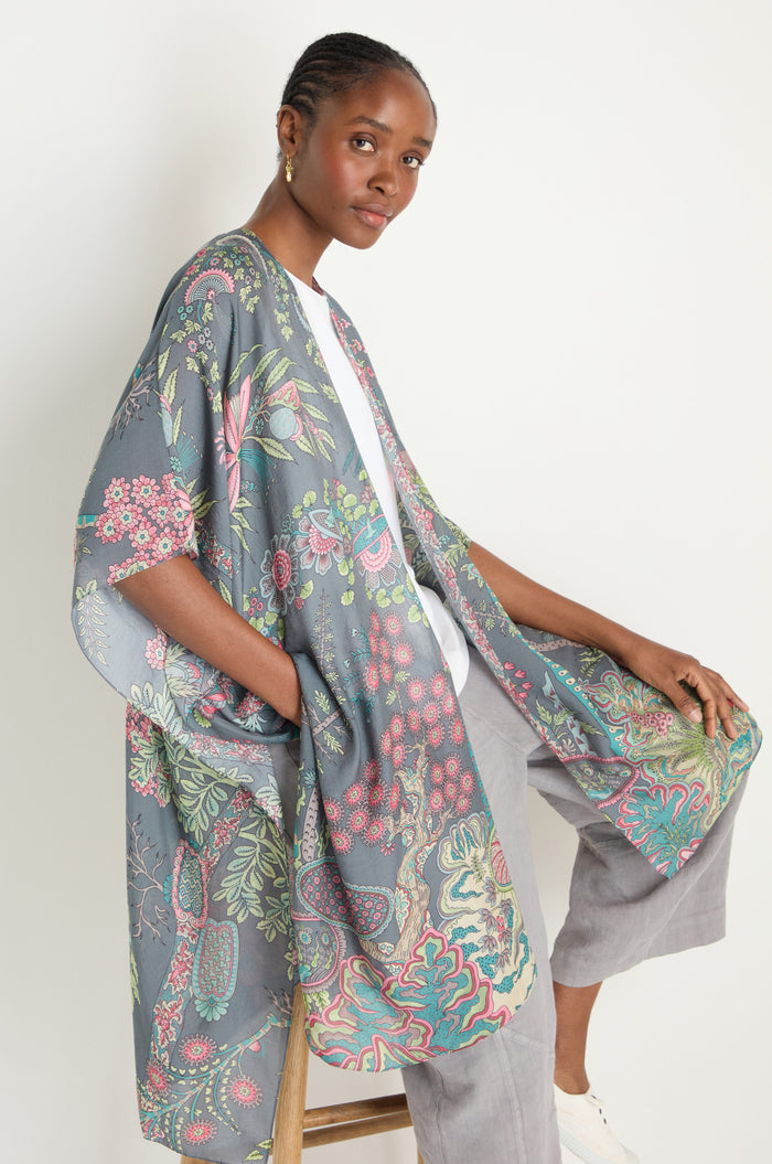 A woman sits on a stool wearing a white top, light gray pants, and the Boho Garden Print Throwover in shades of blue, pink, and green.