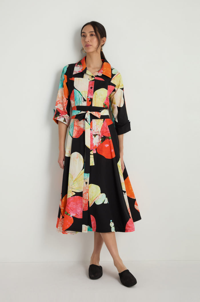 A woman stands against a white wall wearing the Bright Floral Flared Dress featuring long sleeves, a collar, belt, and colorful abstract floral print on black, paired with black slip-on shoes.