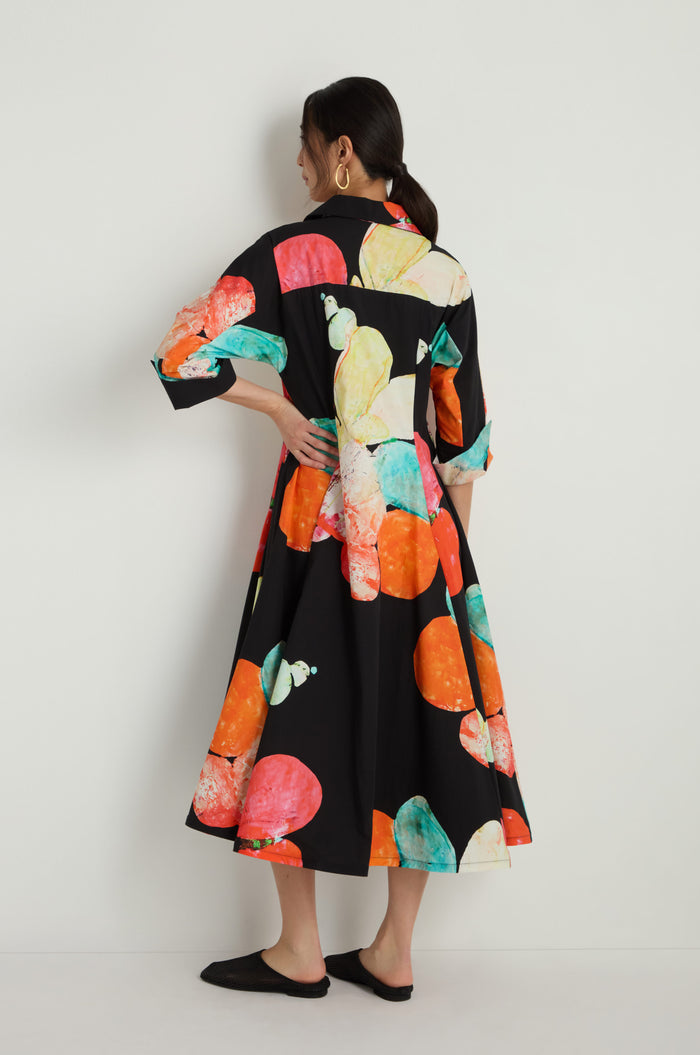 A woman faces away, wearing the Bright Floral Flared Dress—a black cotton midi dress with orange, teal, and yellow abstract circular patterns—standing against a plain white background.