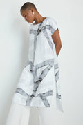 A woman with long gray hair wears the Brushstroke Print Long Tunic, featuring a loose fit and abstract gray patterns. She stands against a plain background, looking down as she adjusts her necklace.