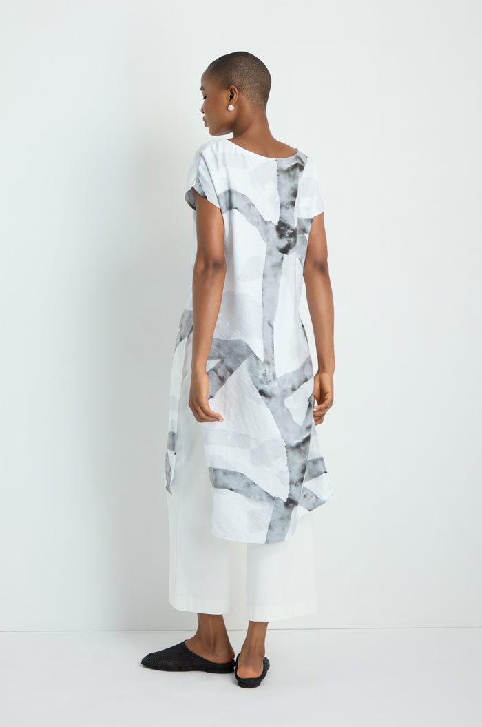A woman with long gray hair wears the Brushstroke Print Long Tunic over black pants and sandals, standing sideways against a plain white background.