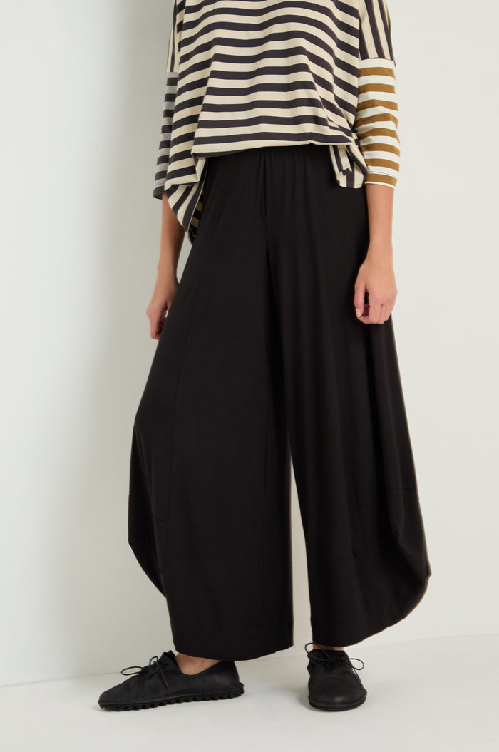 Person wearing a black and beige striped top with asymmetrical sleeves, Bubble Hem Trousers in black, and black shoes, standing against a plain backdrop.