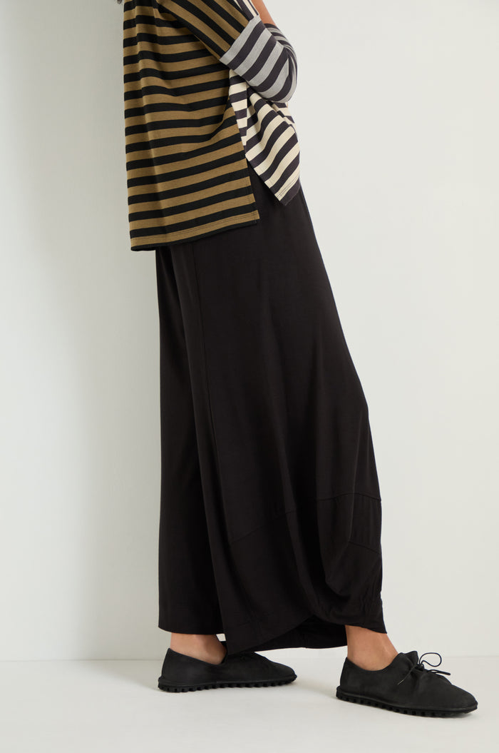Someone wearing Bubble Hem Trousers, a striped long-sleeve top, and black lace-up shoes stands against a plain white wall.
