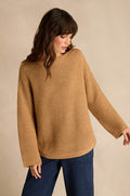 A person wearing the Camel Ribbed Wool Blend Button Jumper stands against a beige background, looking down and to the side. The ribbed wool blend jumper pairs effortlessly with dark blue pants for an understated, classic look.