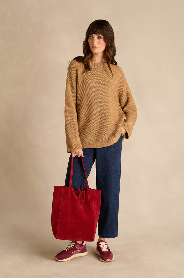 A woman wearing the Camel Ribbed Wool Blend Button Jumper, blue jeans, and red sneakers, holding a large red tote bag, stands against a neutral background.