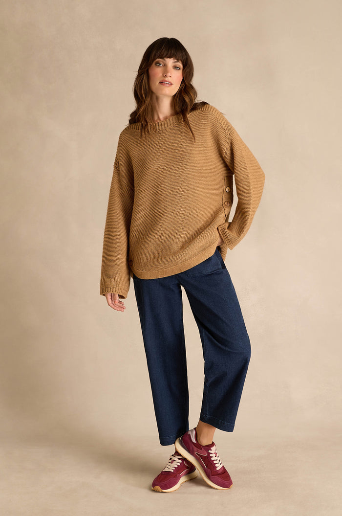 A woman wearing a Camel Ribbed Wool Blend Button Jumper, blue wide-leg pants, and red sneakers stands against a plain beige background.