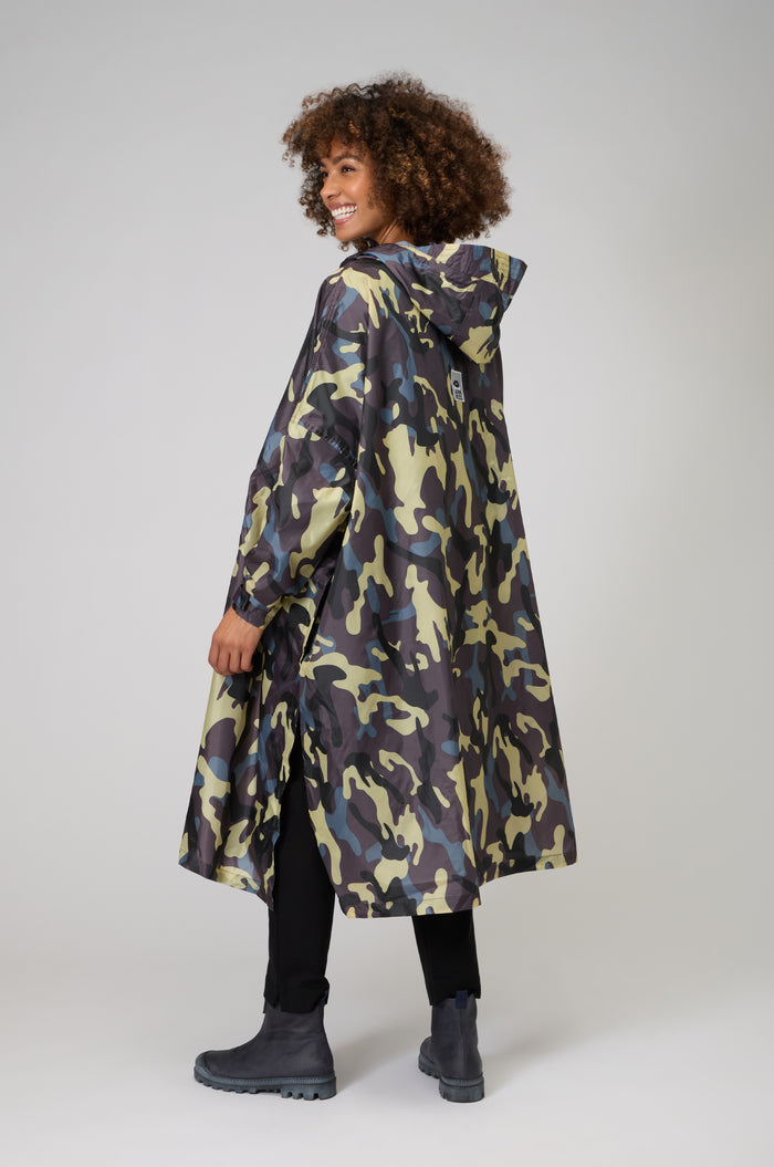 A person stands and smiles against a plain background, wearing the Camo Poncho along with black pants and boots.