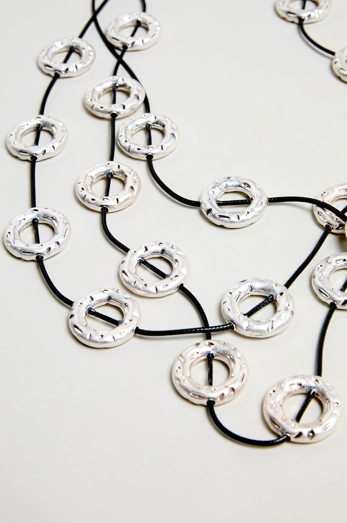 The Cascade Hoops Necklace showcases layered silver hoops with a textured finish, joined by black cords and arranged in overlapping circles against a light background.