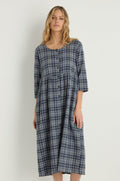 A woman stands against a plain background wearing the Check Button-Through Navy Dress, featuring a blue and white plaid Danish design with three-quarter sleeves.