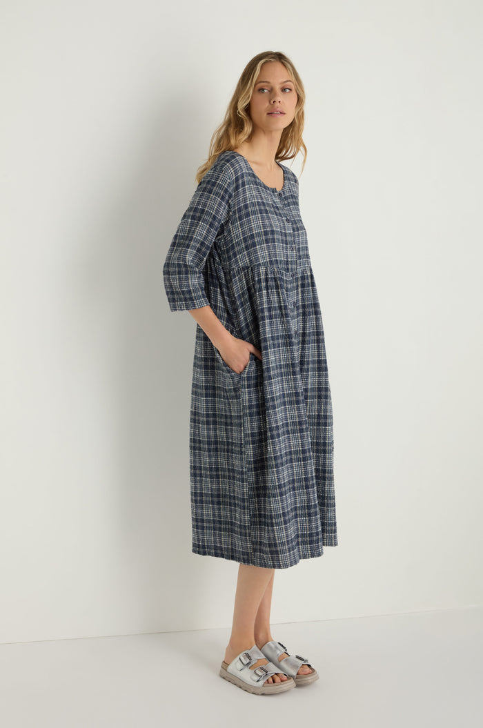 A woman stands against a plain white background wearing the Check Button-Through Navy Dress—a blue plaid, textured cotton dress with three-quarter sleeves—paired with white sandals, reflecting the effortless charm of Danish design.