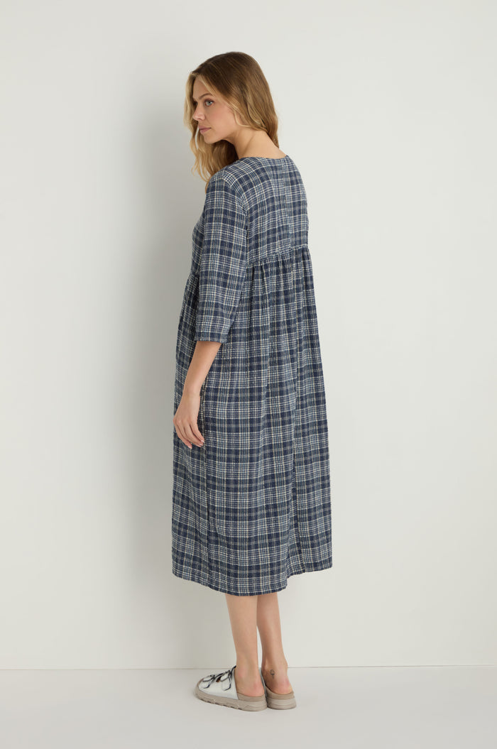 A woman stands sideways in the Check Button-Through Navy Dress, a loose-fitting cotton piece with blue plaid texture and three-quarter sleeves, paired with light sneakers, inspired by Danish design against a plain background.