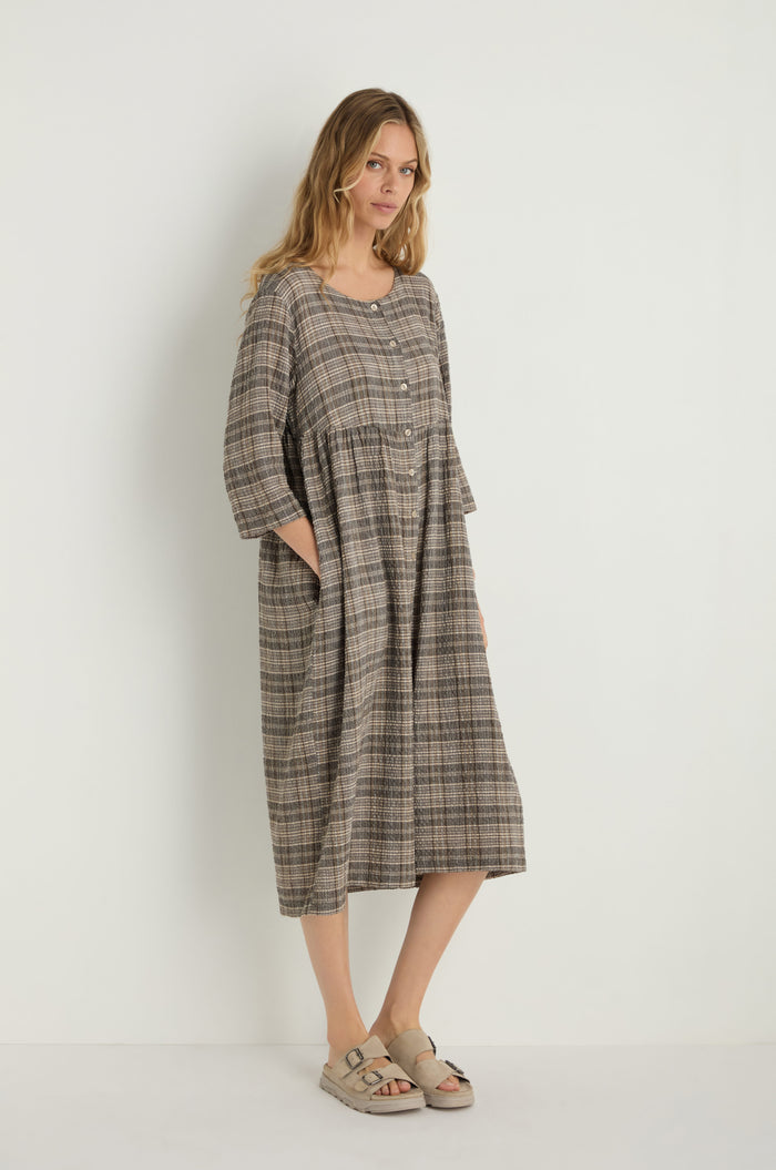 A woman models the Check Button-Through Taupe Dress in plaid textured cotton with three-quarter sleeves and beige sandals, standing against a plain white background.