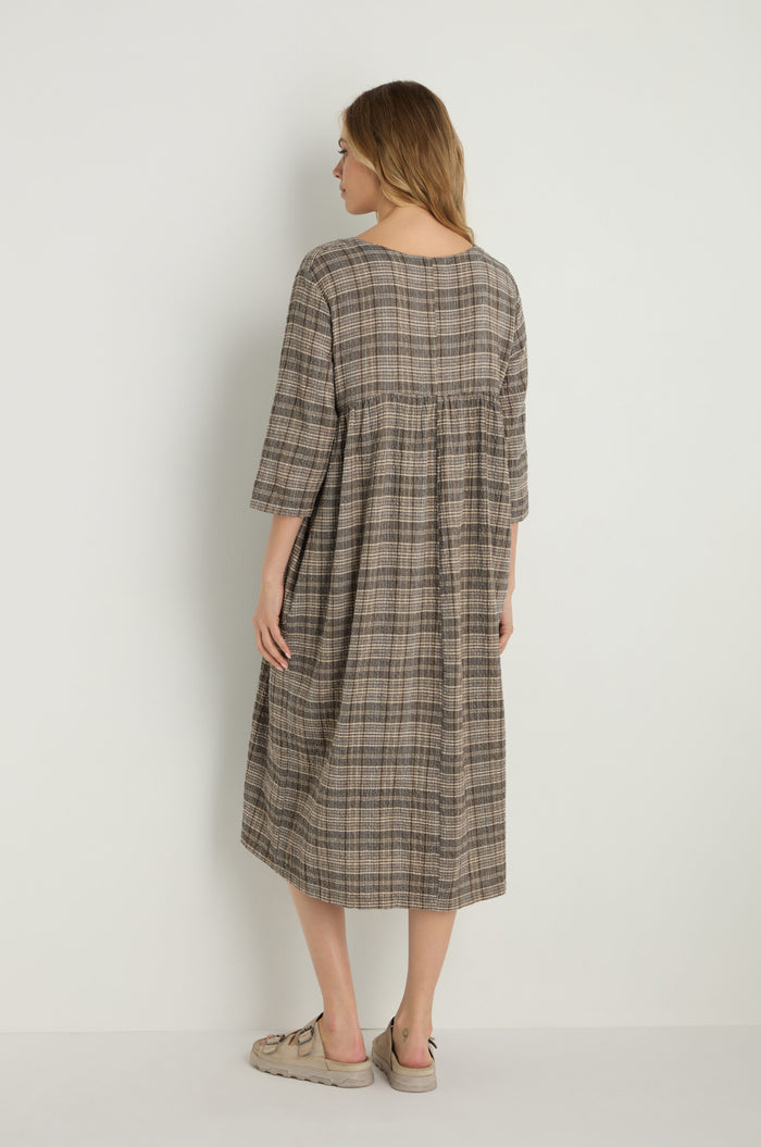 A woman faces away, wearing the Check Button-Through Taupe Dress—a loose-fitting, knee-length plaid dress with three-quarter sleeves—paired with light sandals against a plain background.