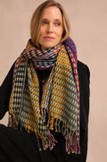 A woman wears the Checkerboard Scarf—an oversized, multicolored accessory—draped over a black coat. Seated with her arms crossed and gazing slightly to the side against a neutral background, she lets the lightweight wool-blend scarf add warmth and a vibrant pop to her ensemble.
