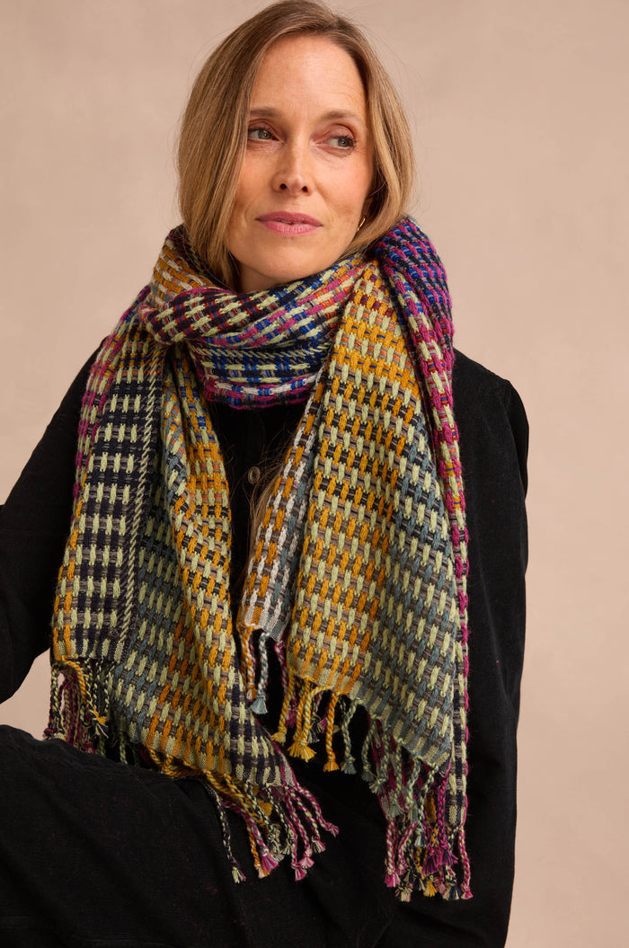 A woman wears the Checkerboard Scarf—an oversized, multicolored accessory—draped over a black coat. Seated with her arms crossed and gazing slightly to the side against a neutral background, she lets the lightweight wool-blend scarf add warmth and a vibrant pop to her ensemble.