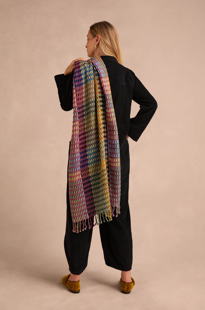 Wearing a black outfit and facing away, the person drapes the Checkerboard Scarf—a colorful wool-blend accessory—over their left shoulder, bringing lightweight warmth to a neutral backdrop.