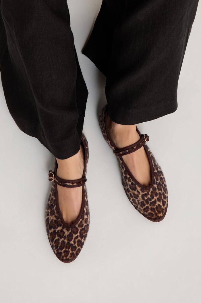 A person stands on a light surface wearing black pants and Cheetah Mary Jane Shoes—handmade in Spain with ankle straps and a recycled bicycle tyre sole.