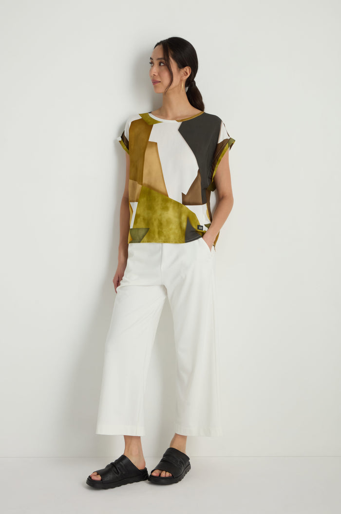 A woman stands against a white wall wearing the Collage Print Top in a relaxed fit, paired with white wide-leg pants and black sandals.