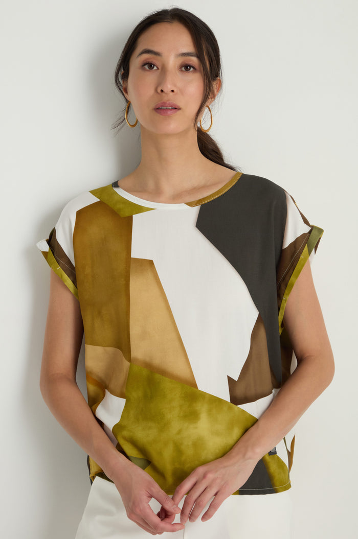 A woman stands against a white wall wearing the Collage Print Top, featuring a relaxed fit, soft viscose fabric, an abstract green, brown, and white geometric design, paired with gold hoop earrings.